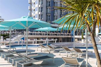 Outdoor pool area with blue umbrellas, lounge chairs, palm trees, and a multi-story building at B&B Hotel Chioggia Airone