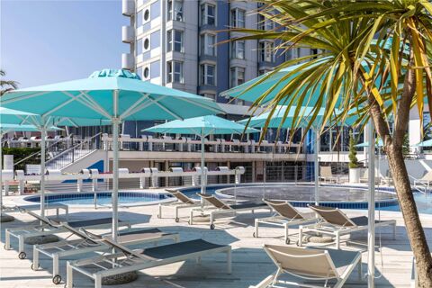 Outdoor pool area with blue umbrellas, lounge chairs, palm trees, and a multi-story building at B&B Hotel Chioggia Airone
