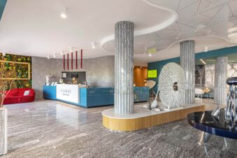 Modern hotel lobby with marble flooring, grey pillars, blue reception desk, red sofa, and decorative art at B&B Hotel Chioggia Airone