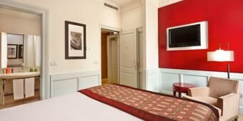 Hotel Indigo Rome-St.George room with bed, patterned bedspread, armchair, red wall panel with mounted TV, and open bathroom door