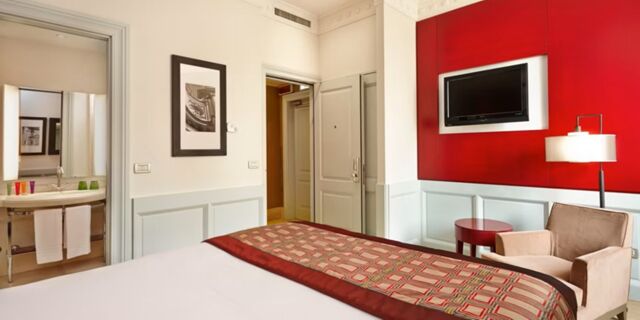 Hotel Indigo Rome-St.George room with bed, patterned bedspread, armchair, red wall panel with mounted TV, and open bathroom door