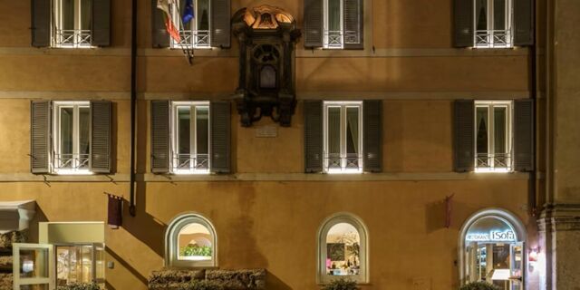 Exterior view of the Hotel Indigo Rome-St.George with illuminated windows and Italian flags against a warm beige wall at night