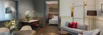 Modern living space in NH Collection Milano Porta Nuova featuring a gray sofa with red cushions, a dining table, and a bedroom visible through an open door