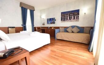 Hotel Raffaello room featuring a bed, sofa with cushions, wooden floor, wall-mounted TV, desk, and blue curtains on the window