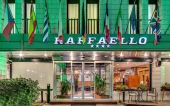 Hotel Raffaello entrance at night with illuminated sign, multiple country flags, glass doors, and outdoor seating visible