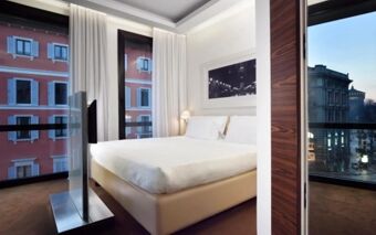 A modern Hotel Una Cusani bedroom with a large bed, white bedding, black and white wall art, and windows showing city buildings