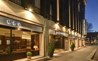 Exterior night view of Hotel Una Cusani entrance with illuminated signage and potted plants along the sidewalk