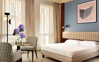 A guest room at Hotel Una Cusani featuring a beige bed with a headboard, two patterned chairs, a round table with purple flowers, and large sheer curtains over windows