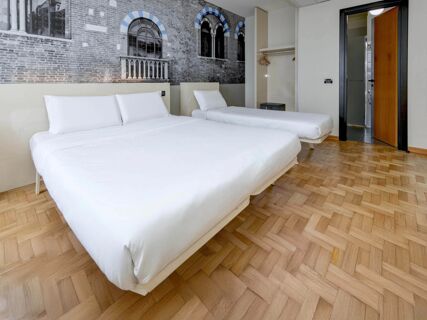 Two neatly made beds with white linens in a hotel room featuring a brick wall mural and wooden parquet floor at B&B Hotel Verona