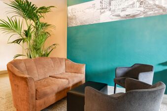 Brown sofa, two gray armchairs, a black ottoman, green plant, and a blue accent wall with artwork at B&B Hotel Verona