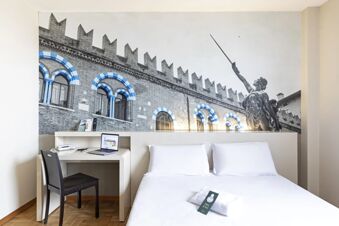 White bed with pillows, desk with laptop and phone, black chair, and a large mural above the bed at B&B Hotel Verona