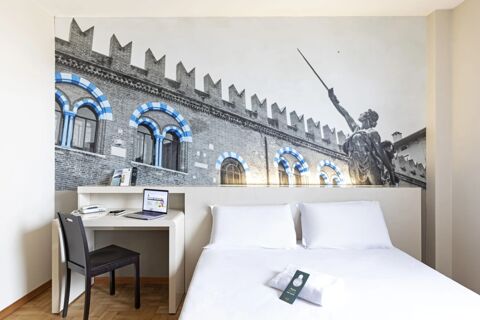 White bed with pillows, desk with laptop and phone, black chair, and a large mural above the bed at B&B Hotel Verona
