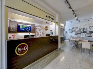 Reception desk with welcome sign above, display screens, breakfast area with tables and chairs at B&B Hotel Verona