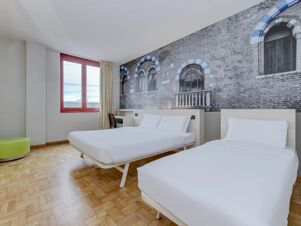 Hotel room at B&B Hotel Verona with two white beds, wooden parquet floor, a red-framed window, and a wall mural of arched windows