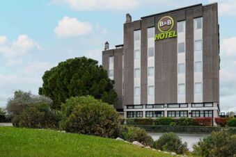 B&B Hotel Verona building with green and white windows surrounded by trees and shrubs under a partly cloudy sky