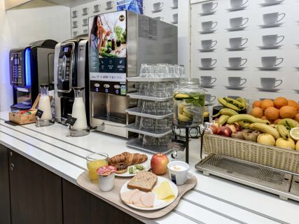 Self-service breakfast area with fruit, juice dispensers, coffee machines, glasses, and a food tray at B&B Hotel Verona