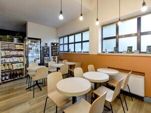 Seating area with light wood chairs and round tables near windows and shelves stocked with snacks in B&B Hotel Verona