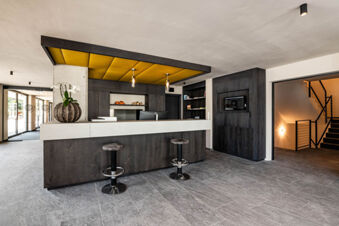 Reception area of Hotel Wochtla Buam with dark wood desk, two stools, yellow ceiling panels, and a staircase in the background