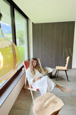Woman in a white robe reading a book while sitting on a lounge chair near a large window at Hotel Wochtla Buam