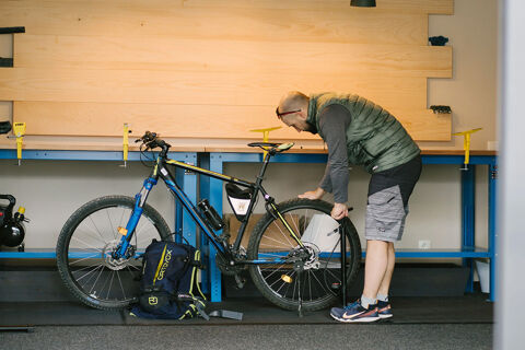 A man inflating a mountain bike tire with a pump in a workshop next to a blue and yellow bike and a backpack at Hotel Wochtla Buam