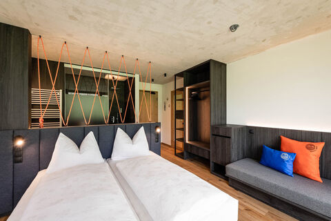 Hotel Wochtla Buam room with two white beds, dark wood furniture, cushioned gray bench with blue and orange pillows, and an open wardrobe