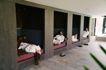Four people in white robes relaxing in wooden alcove seating with cushions at Hotel Wochtla Buam spa area