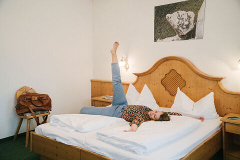A woman lies on her back with one leg raised on a double wooden bed with white bedding at Hotel Wochtla Buam