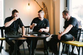 Three men dressed in black uniforms sit around a small table drinking coffee inside Hotel Wochtla Buam