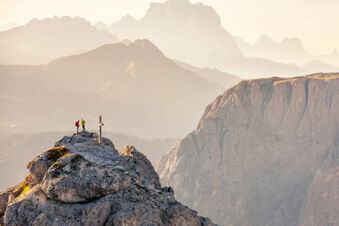 Two hikers stand next to a wooden cross on a rocky summit with layered mountain peaks in the background at Hotel Wochtla Buam