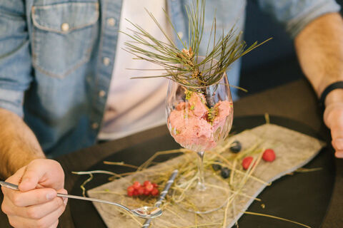 Man holding a spoon beside a glass with pink mousse and pine needles on a wooden board at Hotel Wochtla Buam