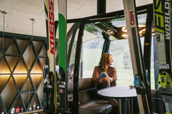 A woman sits barefoot at a round table inside a ski gondola with skis standing outside the windows at Hotel Wochtla Buam