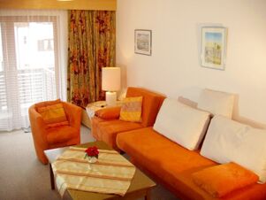 Living room with orange sofa and armchair, coffee table, lamp, floral curtains, and artwork at G&auml;stehaus zur F&auml;rbe Ferien-Appartements