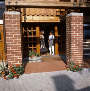Two women stand and talk at the entrance of G&auml;stehaus zur F&auml;rbe Ferien-Appartements with brick pillars and flower pots nearby