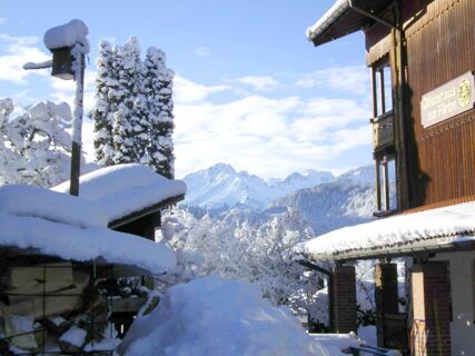 Schneebedeckte Holzh&auml;user und B&auml;ume vor einer Bergkulisse mit Gipfeln im Hintergrund beim G&auml;stehaus zur F&auml;rbe Ferien-Appartements im Winter.