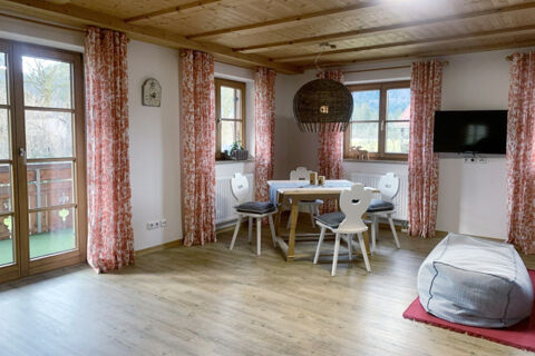 Dining area with wooden table, four white chairs with cushions, large windows with patterned curtains, and a TV at Landhaus Berktold