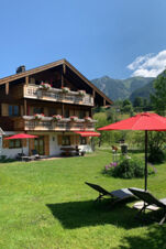 A wooden chalet with flower-adorned balconies and red umbrellas outside on a green lawn at Landhaus Berktold
