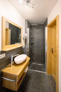 Modern bathroom at Landhaus Berktold featuring dark gray tiles, wooden vanity with white vessel sink, mirror, and glass shower enclosure