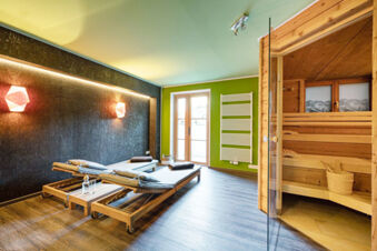 Relaxation room at Landhaus Berktold with wooden lounge chairs, cushions, a sauna with glass door, and ambient lighting on a dark wall