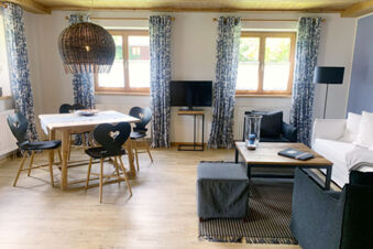 Spacious living room at Landhaus Berktold with wooden dining table, heart-back chairs, white sofa, and patterned curtains on windows