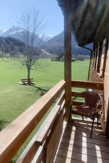 Wooden balcony with a wicker chair at Landhaus Berktold overlooking a green field, a tree, and snow-capped mountains in the distance