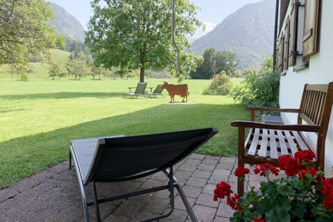 Outdoor seating area with lounge chair, wooden bench, red flowers, green lawn, metal cow sculpture, and mountains at Landhaus Berktold