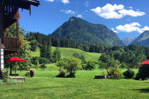 Green lawn with red umbrellas and flowers in front of Landhaus Berktold and forested mountain under blue sky with clouds
