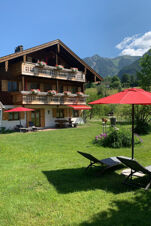 Wooden chalet with flower-decorated balconies and red umbrellas on the lawn at Landhaus Berktold with mountains behind