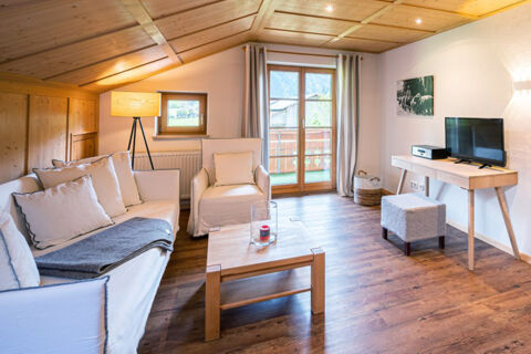Living room at Landhaus Berktold with wooden ceiling and floor, white sofa and armchair, coffee table, TV, and balcony door with curtains