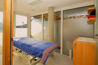 Behandlungsraum mit Massageliege und blauer Decke im Panorama-Hotel Kaserer, umgeben von Fenstern mit Bergmotiven f&uuml;r entspannte Atmosph&auml;re.
