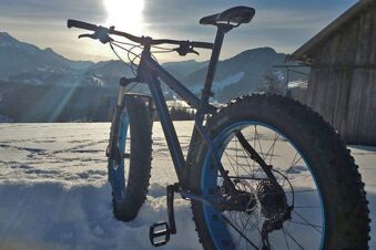 Ein Mountainbike mit dicken Reifen steht im Schnee mit Bergen und Sonnenlicht im Hintergrund, aufgenommen beim Panorama-Hotel Kaserer.