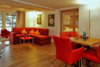 Gem&uuml;tlicher Wohnbereich mit rotem Sofa und St&uuml;hlen sowie Holzboden im Panorama-Hotel Kaserer, inklusive Essbereich und dekorativen Bildern.