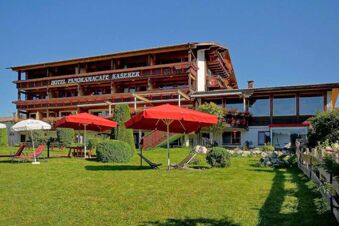 Die Fassade des Panorama-Hotel Kaserer zeigt ein traditionelles Geb&auml;ude mit Balkonen und einer Terrasse mit roten Sonnenschirmen auf einer gr&uuml;nen Wiese.