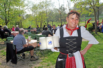 A smiling waitress in traditional dress holds several large beer mugs at outdoor tables of Landgasthof Hotel Hammerm&uuml;hle with customers seated on green chairs and trees in the background