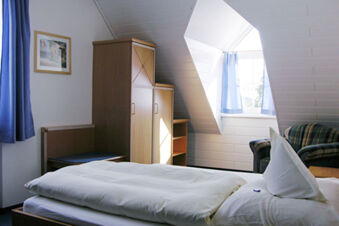 A neatly made bed with white linens next to a wooden wardrobe, blue curtains, and a plaid armchair in Landgasthof Hotel Hammerm&uuml;hle bedroom